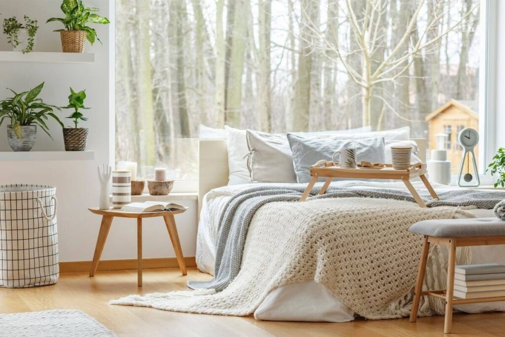 an emotional escape room decorated in minimalist style and pale neutral colors with a bed small side table large window snack tray green plants crocheted blanket several fluffy pillows and books for someone to relax and enjoy some quality mental health time