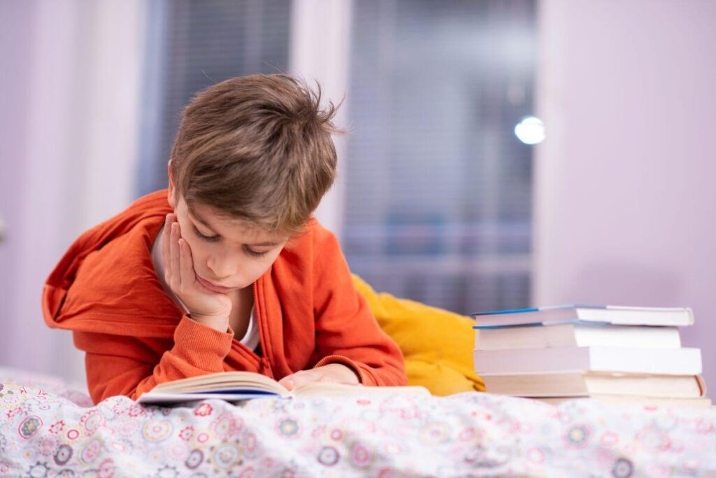 a young boy lounges on a bed and reads a book in a quiet space as he calms down and relaxes