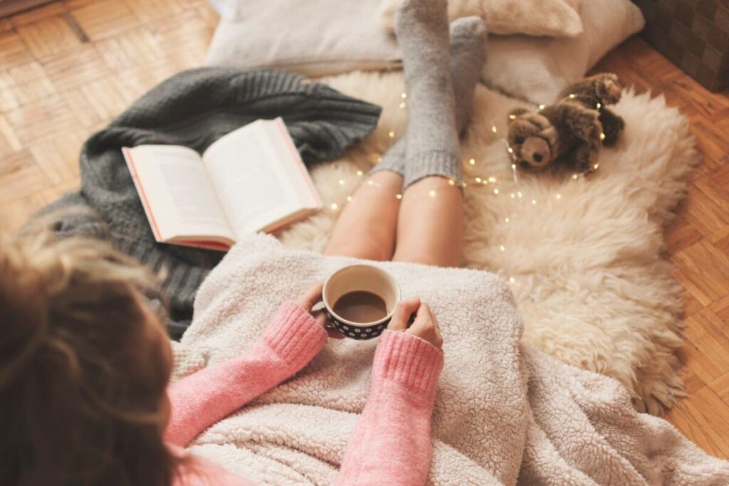 a woman sites on a fuzzy rug on the floor with a blanket over her with fairy lights and a teddy bear and a sweater and pillows and a book holding a cup of coffee relaxing in an emotional escape space