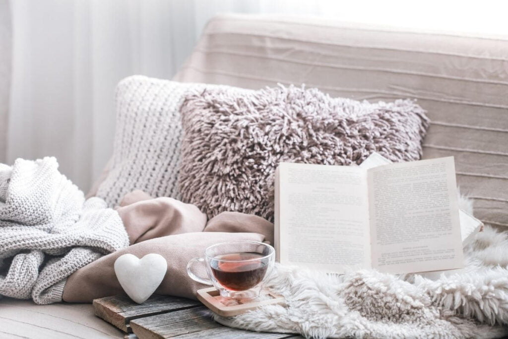 two fuzzy pillows on a soft chair with a book and blanket and sweater with a cup of tea in front of a window for someone to have a comfy emotional escape space in front of a window