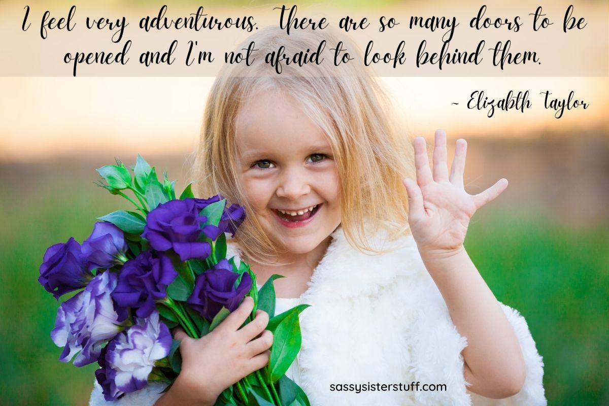 little blonde girl waving at the camera holding a bouquet of purple flowers with a big smile on her face