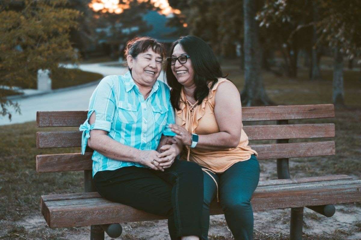 two women sit on a bench hugging each other and laughing