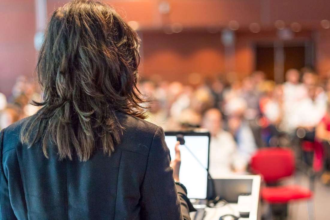 woman standing on stage speaking to an audience