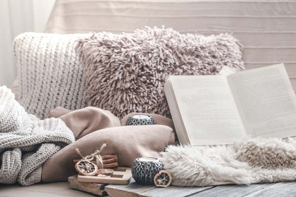 a cozy nook with a book pillows candles blankets in neutral cream colors depicts how to bring more cozy into your life