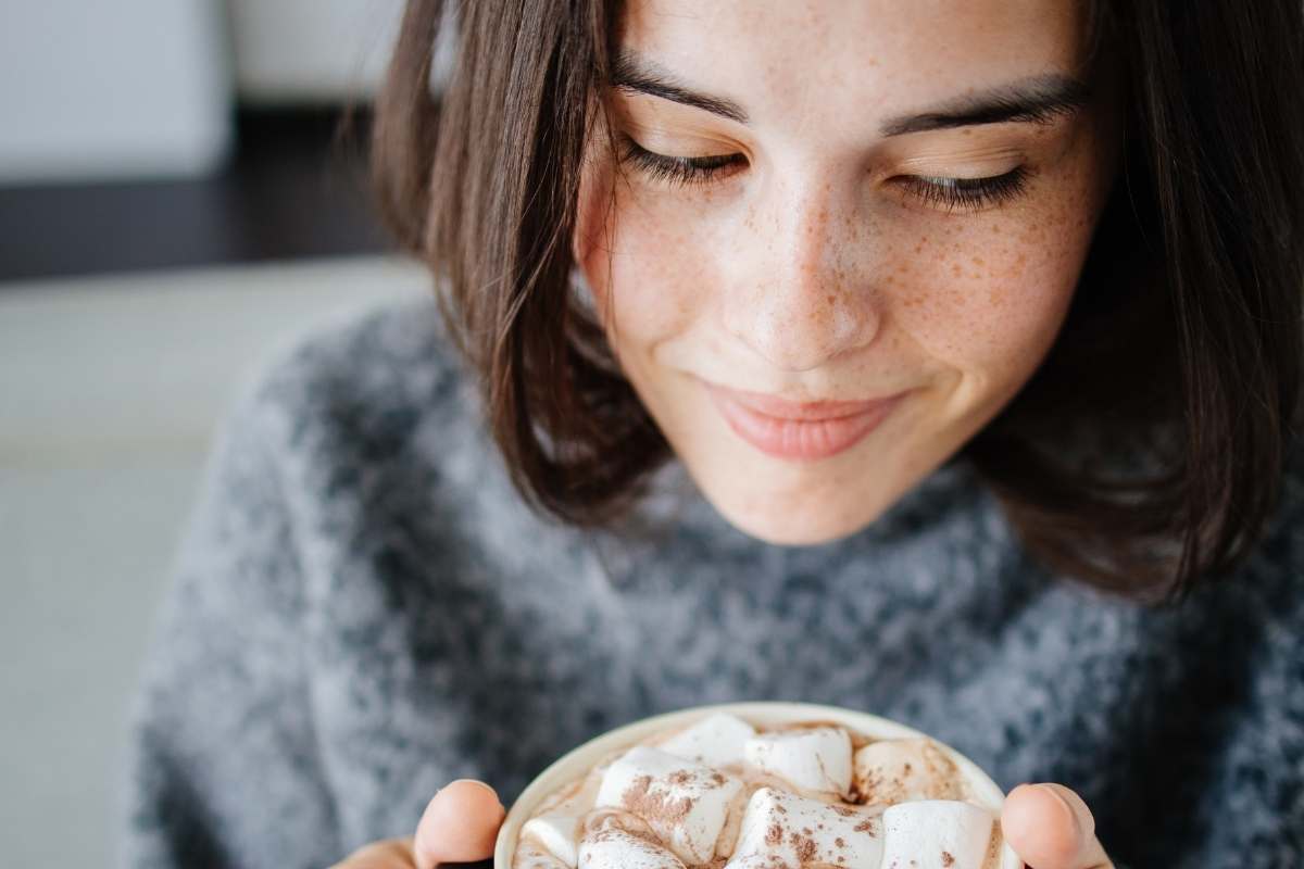 a closeup of a women with freckles smiling and drinking hot chocolate with marshmallows on top