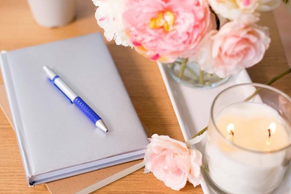 gratitude journal pen candle and pink flowers sit on a light wood table top