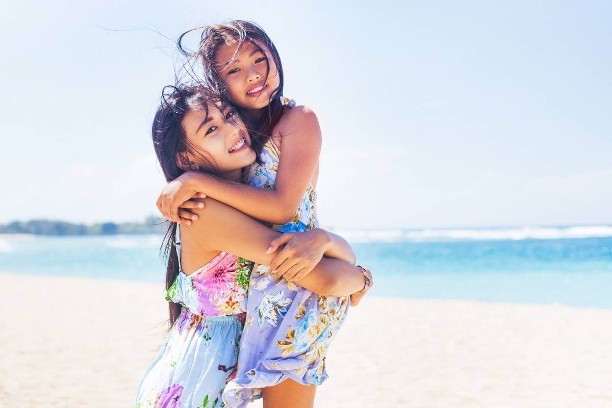 a older girl holding a younger girl on the beach with blue water in the background