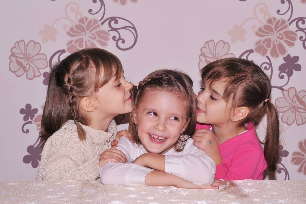 two little girls kissing the head of a third little girl who is smiling real big