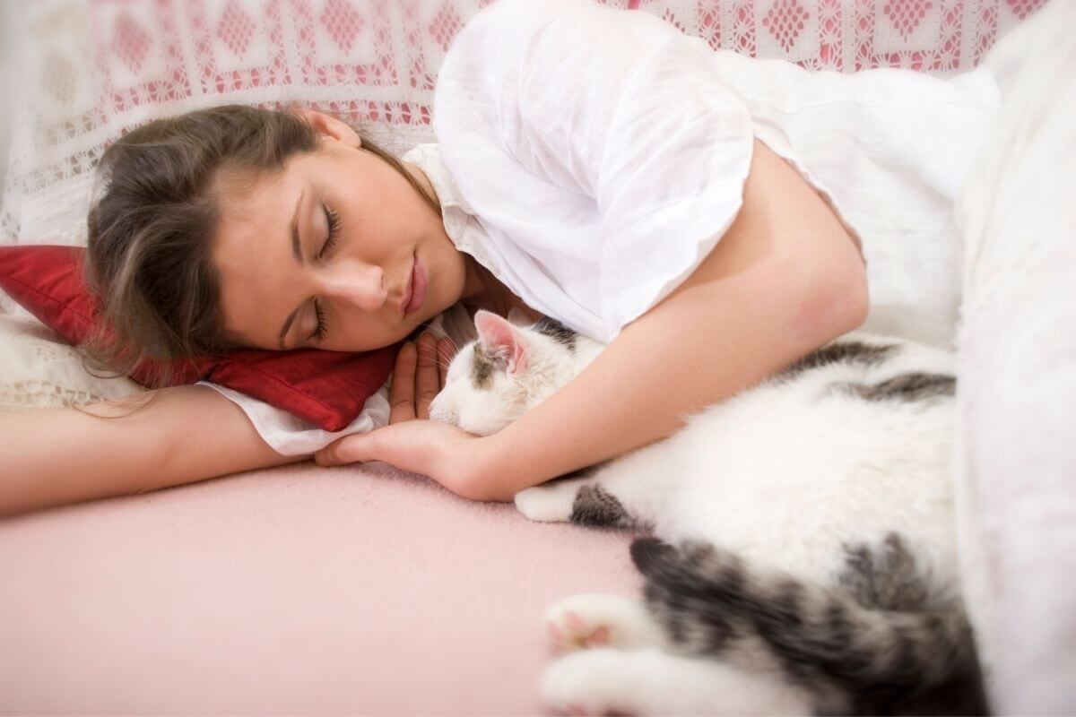 woman sleeping in a white shirt with a white cat tucked under her arm after saying aloud her positive affirmations for good sleep