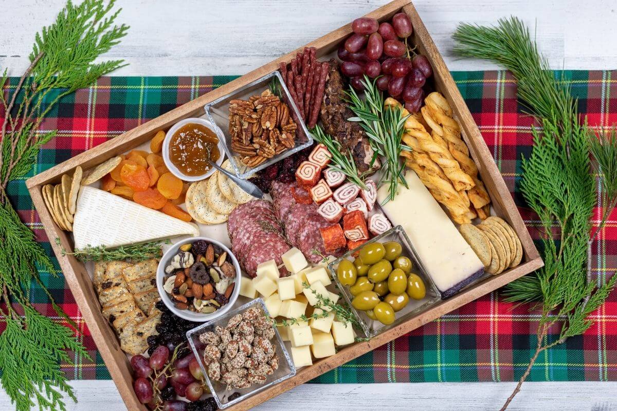 rectangle holiday charcuterie tray with variety of snack foods on a green and red plaid placemat with fresh greenery to decorate