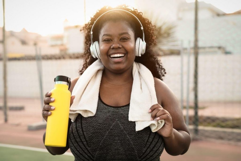 a very happy woman smiles at the camera with headphones a towel and water bottle after exercising