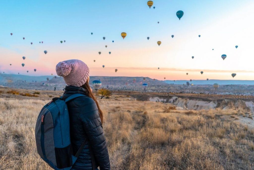 traveler with a backpack on is standing on a hillside looking at dozens of hot air balloons in the sky