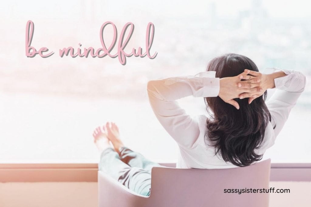 young woman sitting with her hands clasped behind her head looking out a window as she relaxes in mindfulness