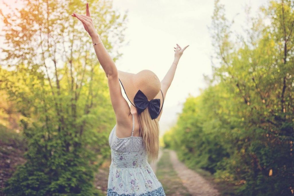 a woman tosses her hands up in the air with happiness as she stands among green trees and thinks about the beauty in life