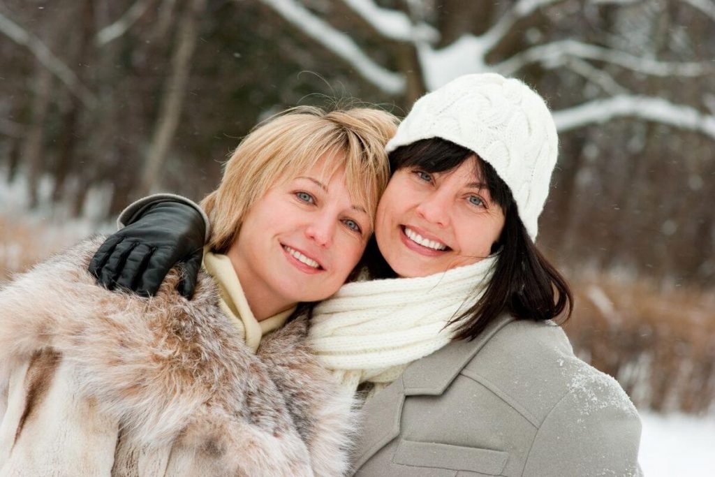 two women in winter coats hugging in a snowy forest as they talk about the beauty of life