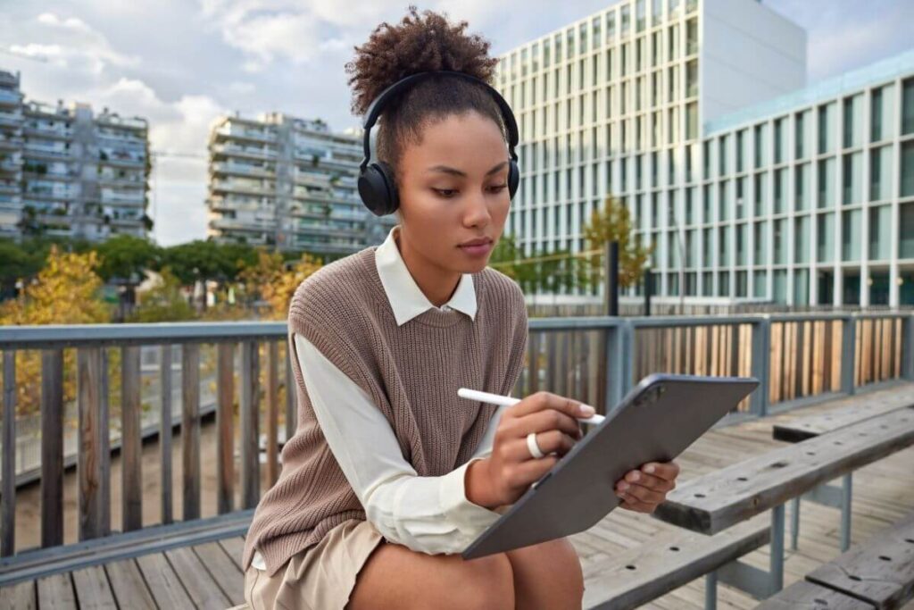 young beautiful woman sits outside on a bench in the city with headphones on working on a table looking very busy as if she may be addicted to stress