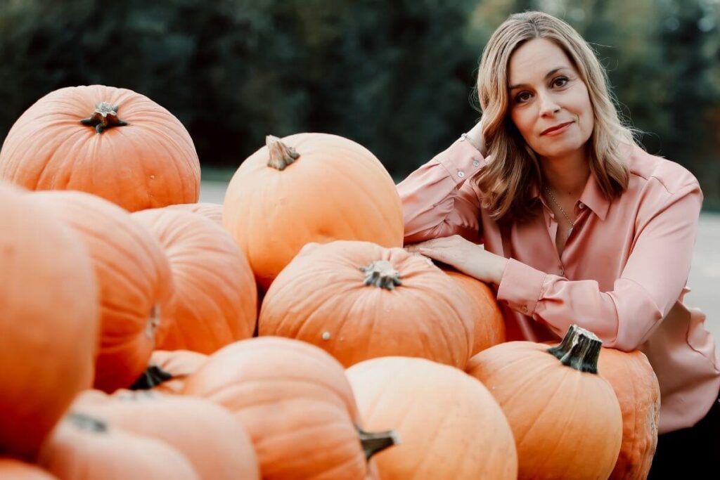 woman with shoulder length blonde hair stands next to a pile of dozens of pumpkins as if wondering how shes going to get these pumpkins where they need to be and why she said YES to this project