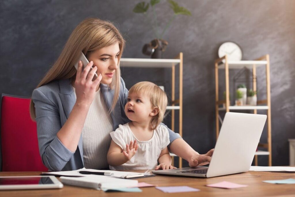 busy woman working from home with her baby sitting in her lap as she multitasks on the phone and working on the laptop in front of her as if possibly addicted to stress and overwhelm