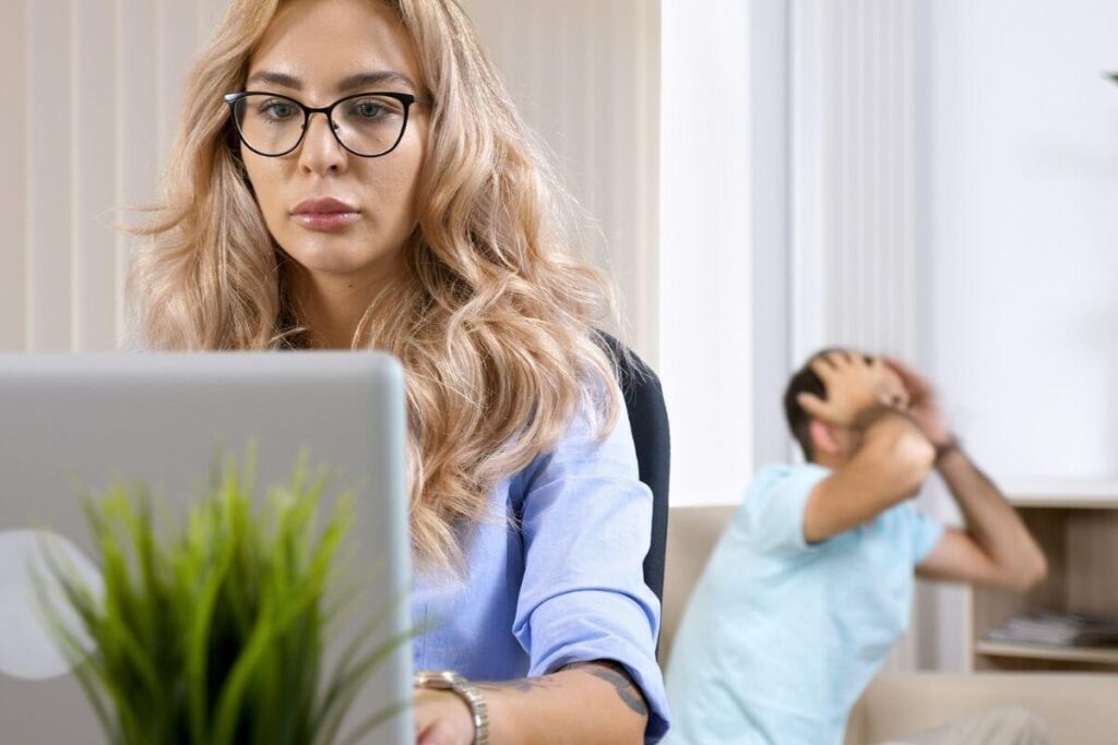 blonde haired woman with glasses sits at a laptop working with a man in a chair behind her clutching his head as if frustrated because hes overwhelmed