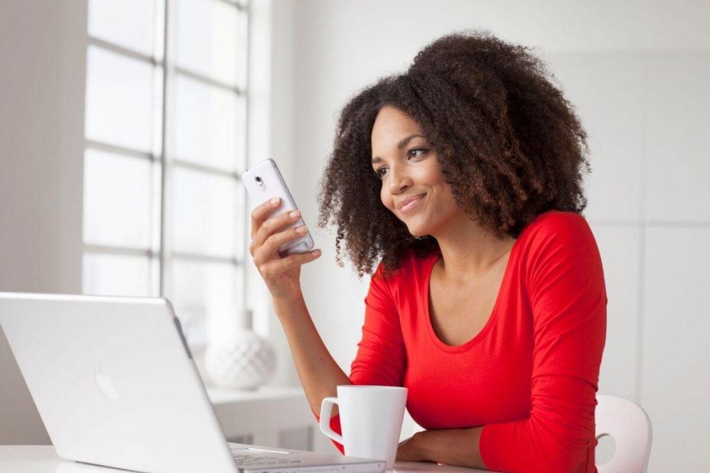 beautiful woman in a red long-sleeved top sits at a desk looking at her phone and laptop at boutique clothes women love