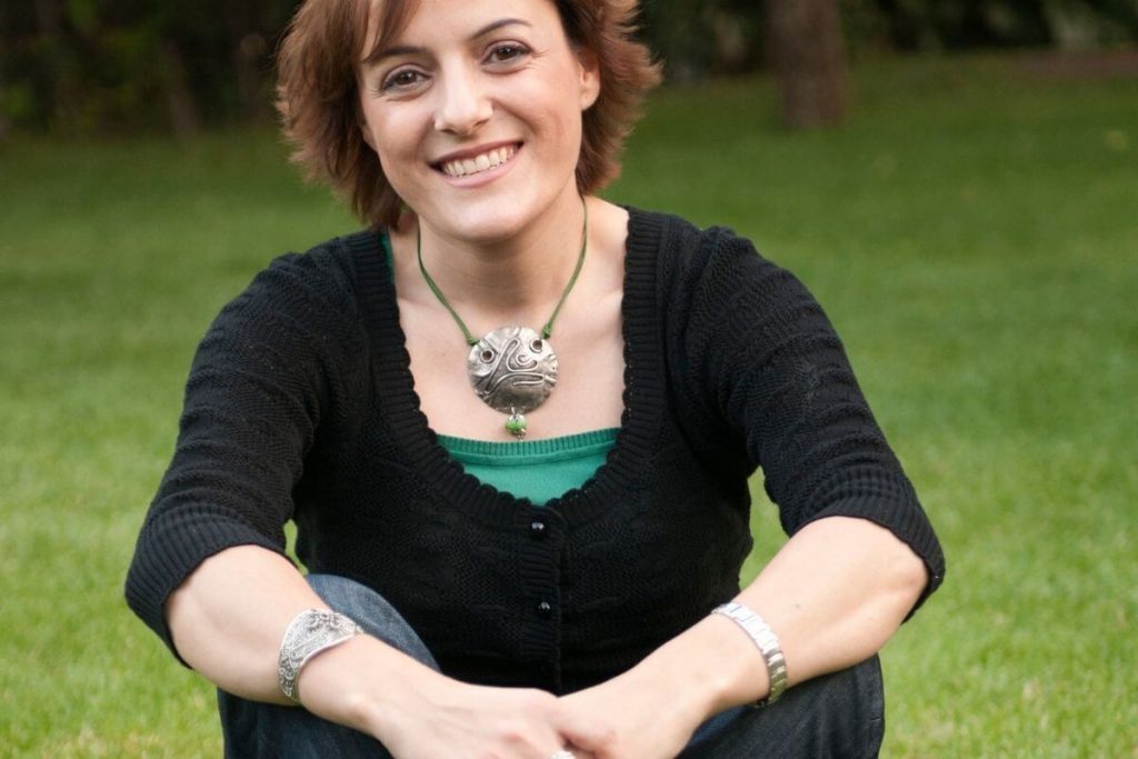 happy women sits on a green lawn wearing a beautiful and trendy large pendant necklace green tank top and black stylish sweater as she smiles at the camera