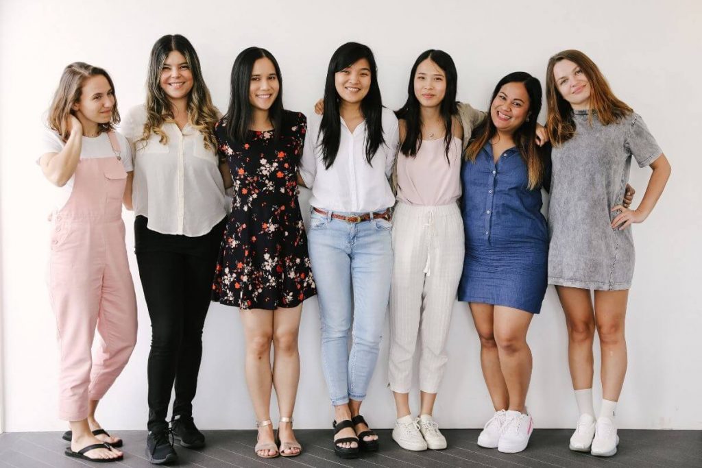 seven young women of varying shapes sizes and styles stand together confidently with their arms around each other smiling at the camera