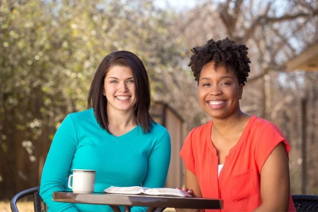 two females study together in an outside wooded setting and smile at the camera confidently