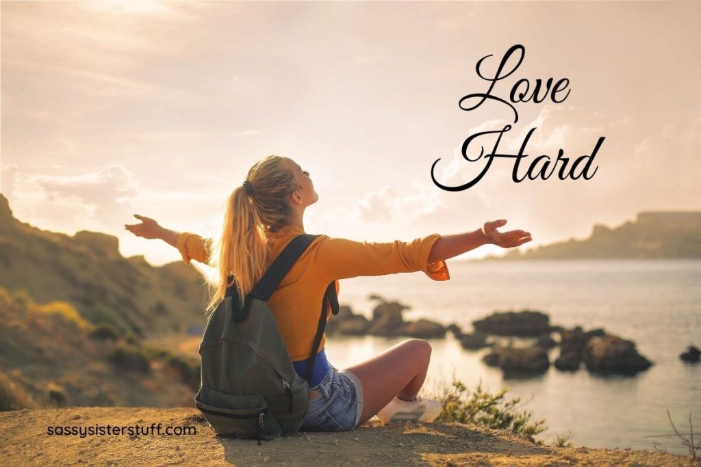 a happy women with a backpack sits on a cliff overlooking a body of water and hills with her hands spread out demonstrating that she is happy and has a healthy mindset