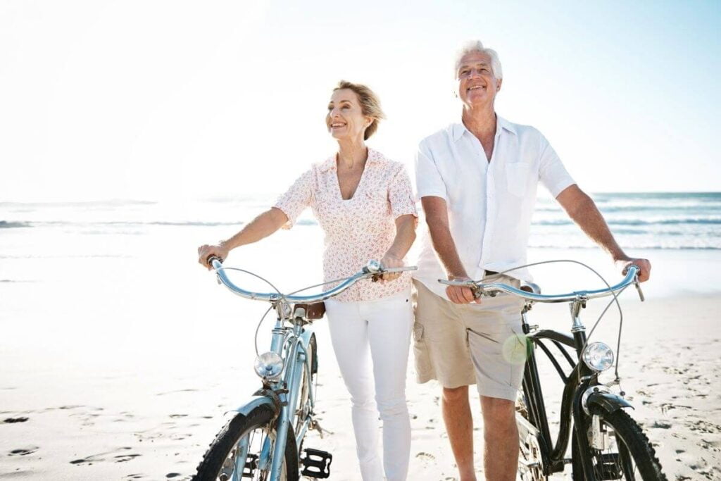 older happy couple in ivory and white casual clothes stand next to their bikes smiling on a beach showing us how to live your best life