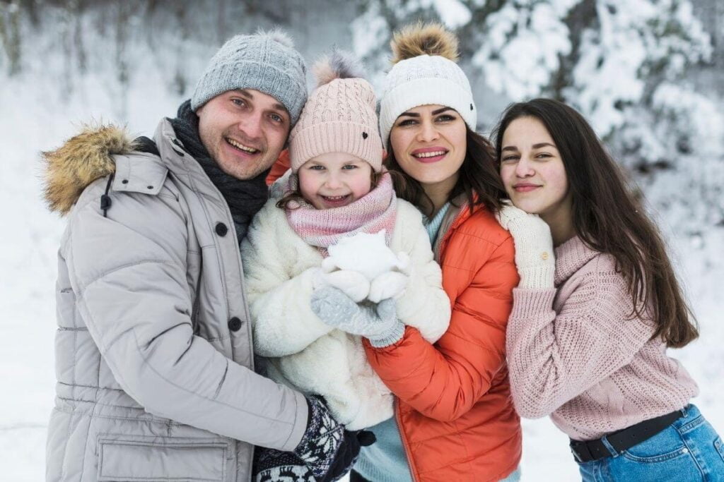 a mom dad teenage daughter and little daughter in winter clothes pose for a winter picture in front of snow covered trees