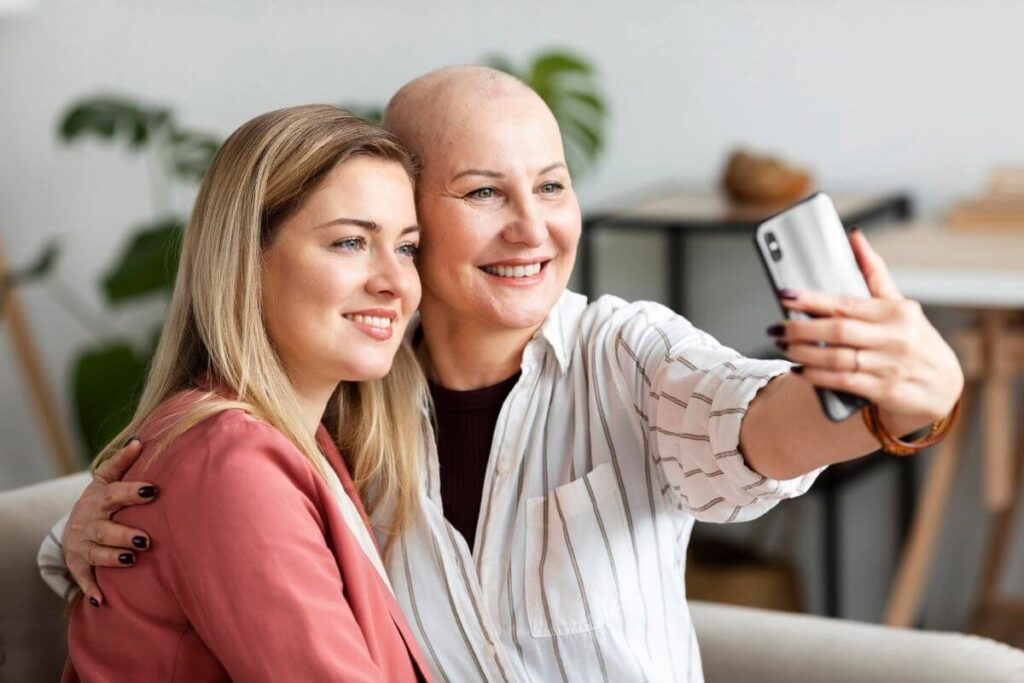 a bald lady and her friend sit on a sofa together and take a selfie