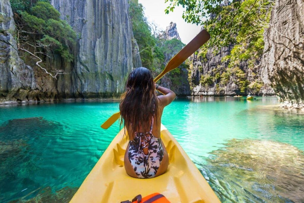 a young woman kayaks among tall rocks and trees in a green body of water in a yellow kayak showing us how to live your best life