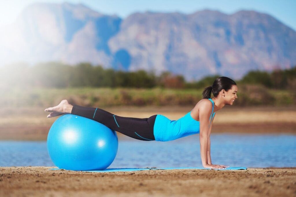 young happy woman exercises with a large ball on a yoga mat in front of mountains and along side a lake