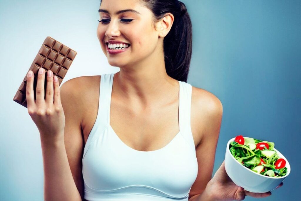 a young happy woman in a white exercise top holds a bar of chocolate in one hand and a bowl of salad in the other smiling as she shows a life of balance and how to live your best life