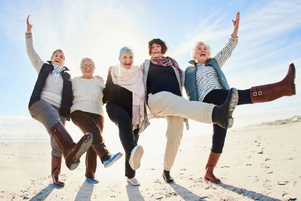 5 happy middle aged and older women in casual clothes walk along a beach kicking up with their legs having fun showing us how to live your best life