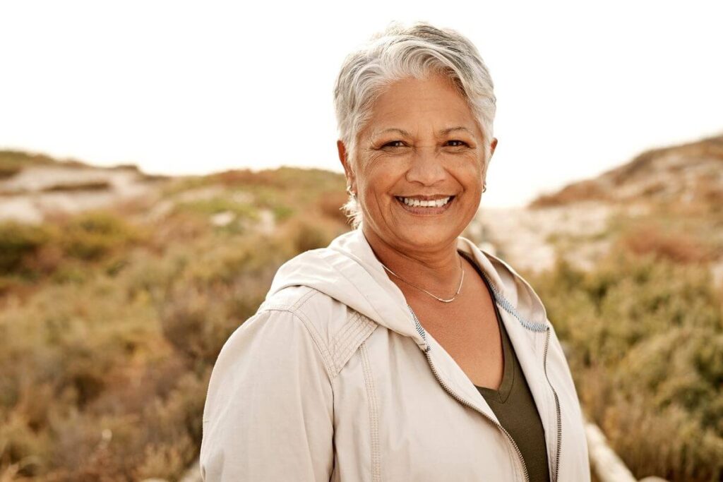 a middle aged woman with short gray hair smiles at the camera as she hikes through hills wearing an ivory jacket showing us how to live your best life