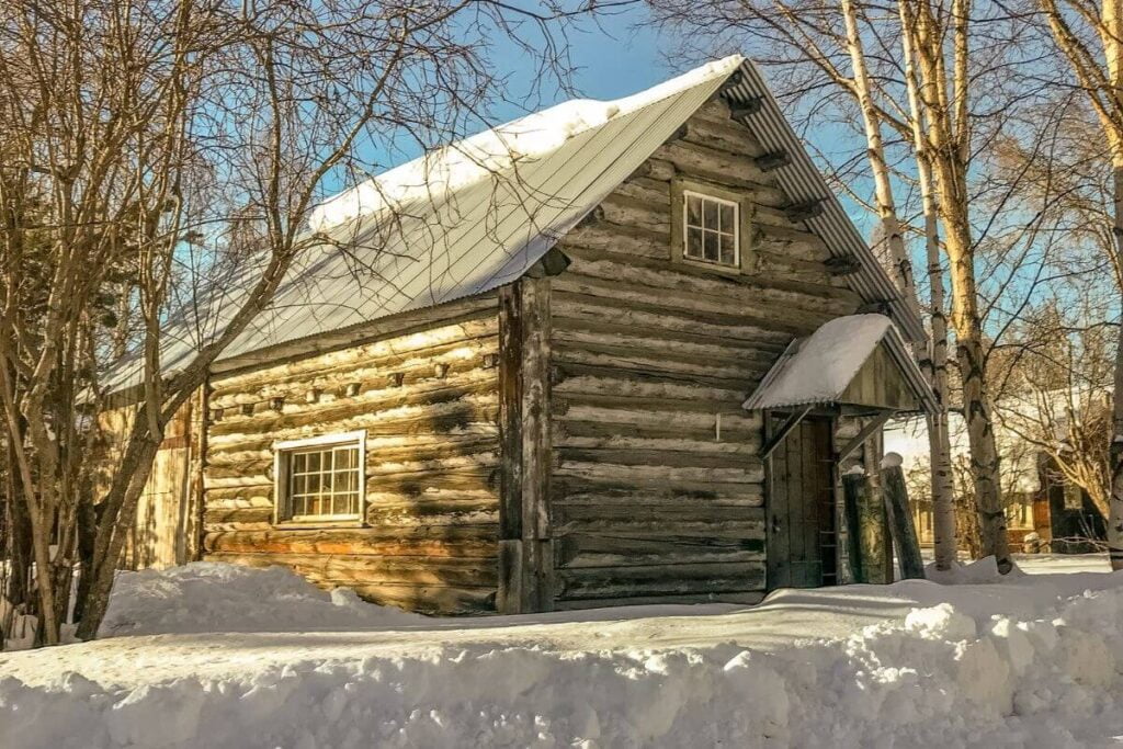 an old log cabin sits in a snow covered area with bare trees around it