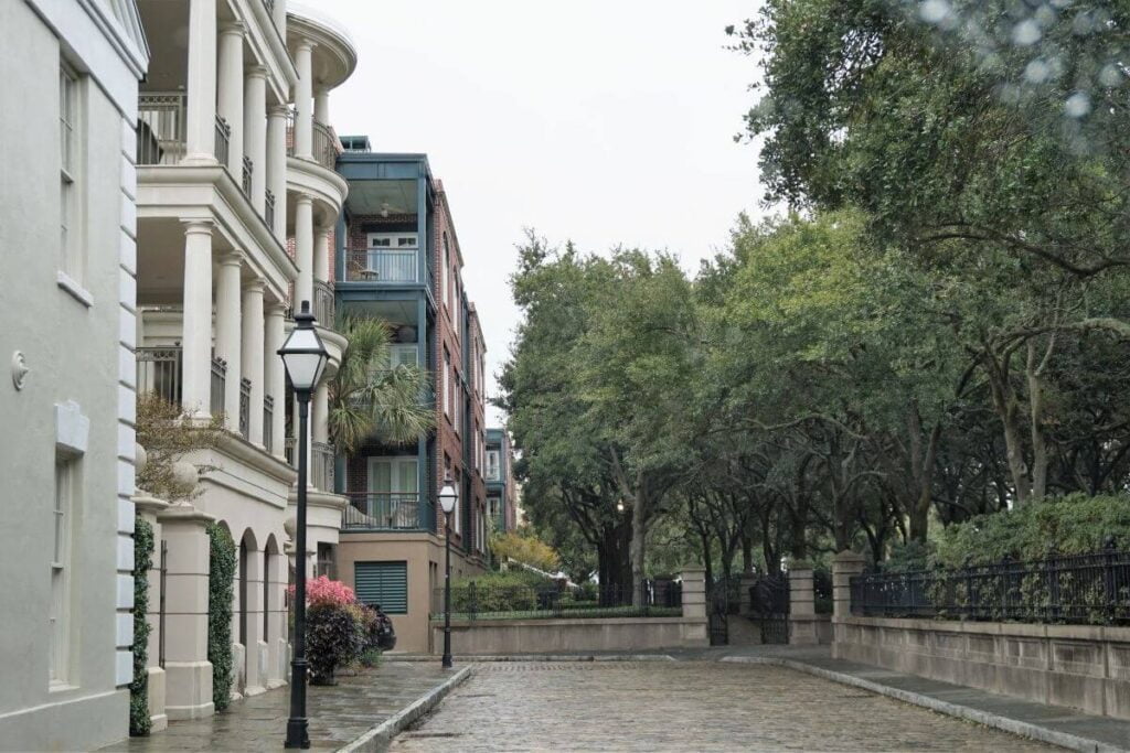 cobble stone street leading to the entrance of the waterfront park in charleston sc with buildings on one side and green trees on the other side of the street