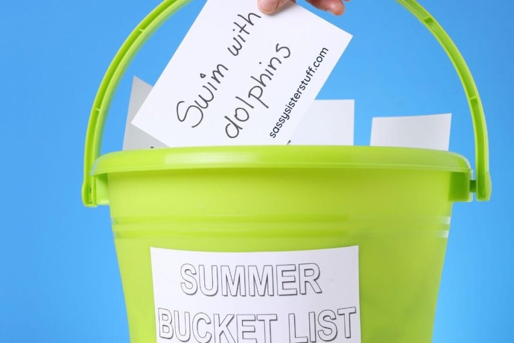 blue background with bright green bucket and white cards with bucket list ideas inside the bucket and a white card that says summer bucket list on the outside and someone picking out a card that says swim with dolphins