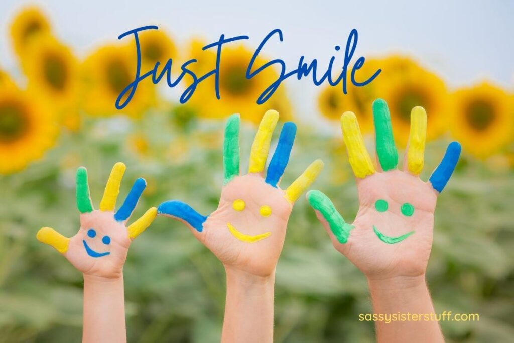 three childrens hands with happy faces painted on their palms in blue yellow and green in front of a field of sunflowers and a message that say just smile