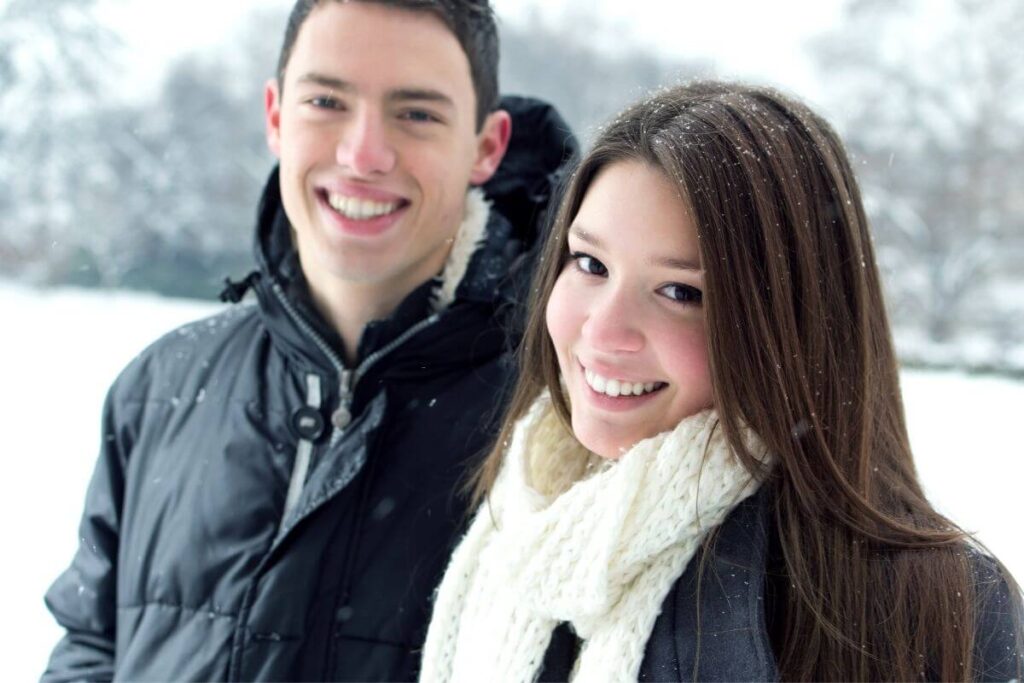 a young woman and young man smile super big at the camera even in a cold snow covered field