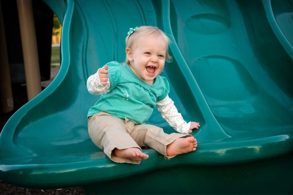 baby girl sliding down a green sliding board at the park with a huge smile on her face