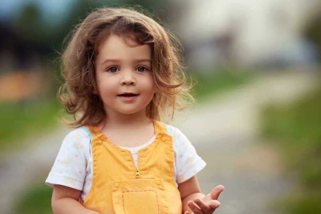 an adorable female toddler with messy brown hair and yellow overalls smiling at the camera