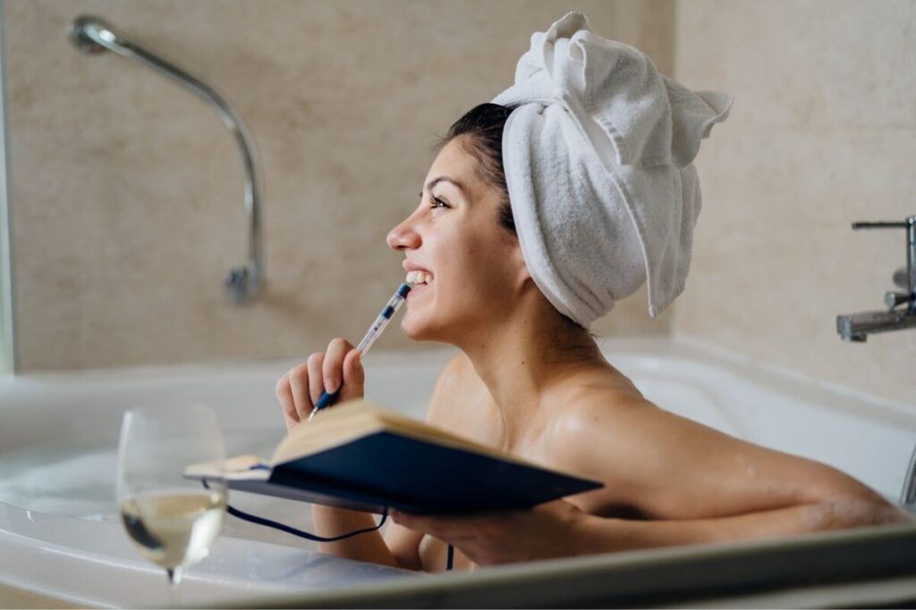 beautiful happy young woman sits in a bathtub with her hair in a towel sipping a glass of wine and writing in her gratitude journal for a self-care challenge