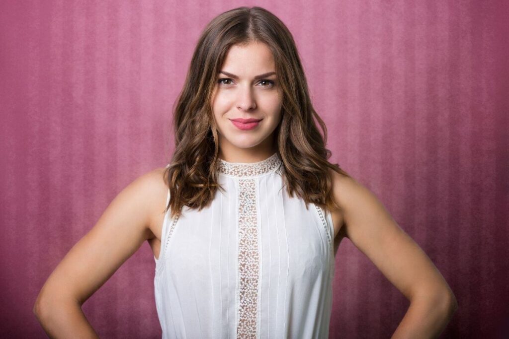 beautiful young woman stands in a white dress with her arms on her hips against a burgundy backdrop looking confidently into the camera and smiling