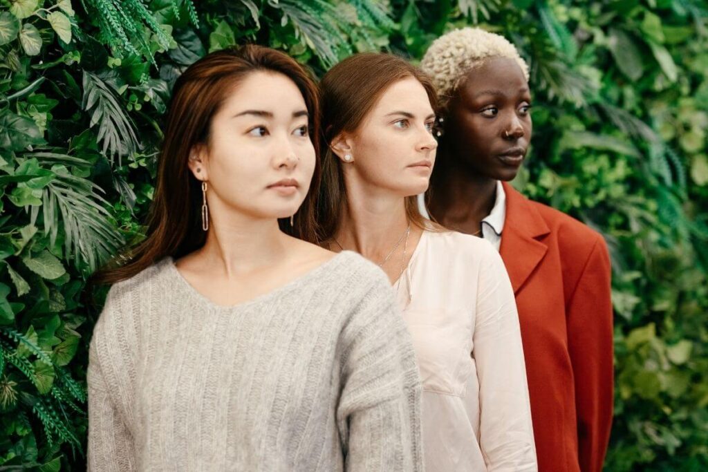 an asian woman a white woman and a dark skinned woman stand in a row outside in front of greenery wearing business attire and looking off into the distance to demonstrate their confidence