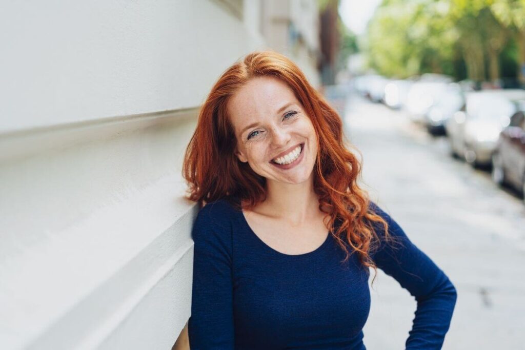 a middle-aged woman with red hair stands on a tree lined sidewalk leaning against a white wall smiling big and looking at the camera demonstrating that she has found her confidence with a series of affirmations for confidence