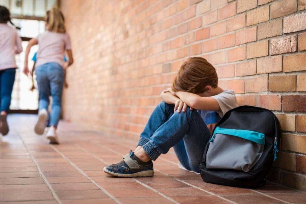 a young boy sits on the floor in a school hallway with brick walls as other children run out to recess without him