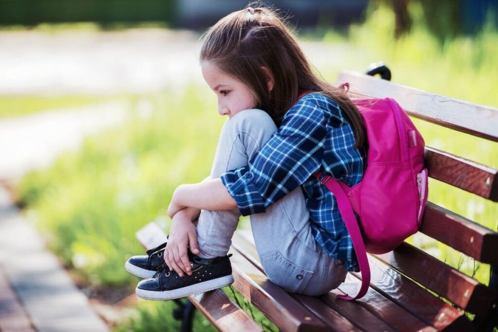 little girl sits on a bench with her backpack on after school looking very sad because the anti-bullying policies at her school are not working and she is being bullies