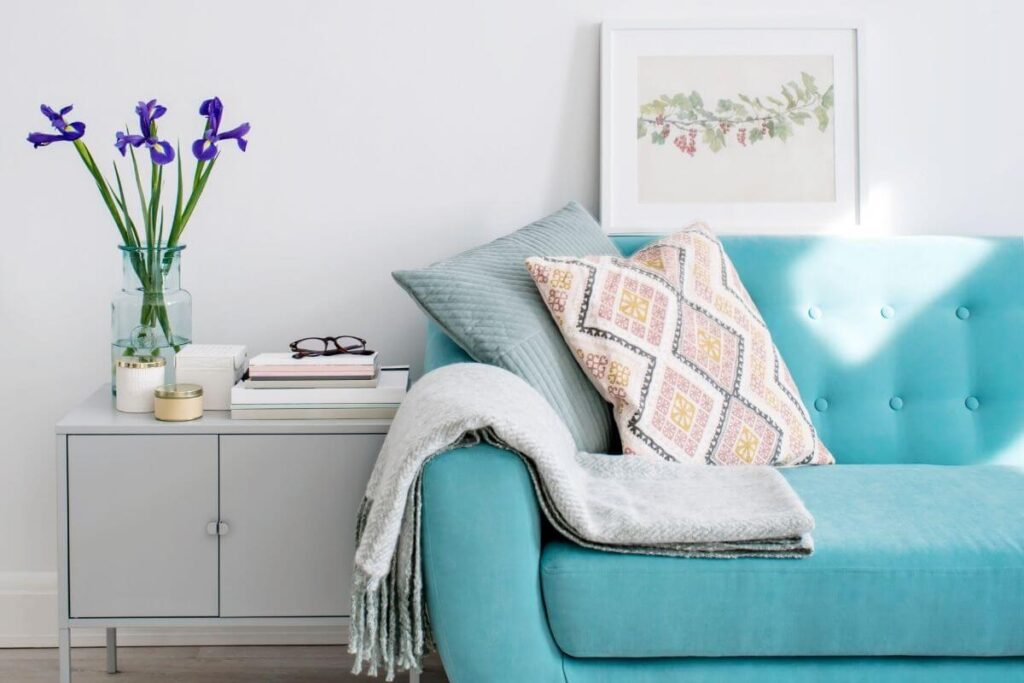 an emotional escape room decorated in shades of gray and white where a turquoise loveseat sits with soft pillows and a floral photo above it and a blanket laying on the arm with candles flower paper books and journals on a simple side table